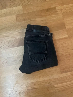 Svarta skinny jeans med slitningar - Säljer ett par svarta skinny jeans med lätt slitna detaljer på knäna. Jeansen har klassisk femficksdesign och normalhög midja. Materialet är stretchigt denim som sitter tajt längs benen för en snygg siluett.
