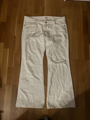 Vita bootcut jeans med slitningar - Säljer ett par vita bootcut jeans med slitna detaljer på låret och knät. Jeansen har fem fickor, knappgylf och broderade bakfickor. Perfekta för dig som gillar en avslappnad och trendig stil.