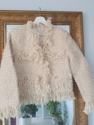 Beige bouclé cardigan med fransar - Ny oanvänd trendig beige bouclé cardiganjacka med fransar längs kanter och nederkant. Jackan har dekorativa guldknappar framtill och två fickor på bröstet. Materialet är strukturerat och ger en lyxig känsla.