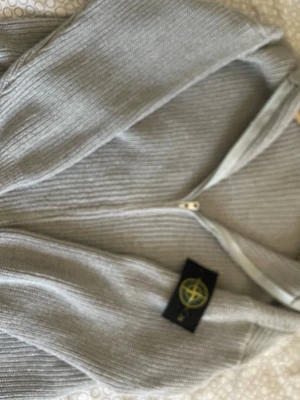 Ljusgrå stickad tröja från med dragkedja - Ljusgrå stickad tröja från Stone Island med dragkedja framtill och ribbad struktur. Tröjan har en klassisk passform och den ikoniska Stone Island-loggan på ärmen. Perfekt för lager-på-lager och enkel att matcha med olika outfits.
