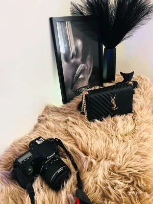 Svart YSL handväskamed guldkedja - Snygg svart handväska från YSL med quiltat mönster och guldfärgad kedjerem. Väskan har ett elegant YSL-emblem framtill och en stilren, rektangulär form. Perfekt accessoar för att lyfta vilken outfit som helst. Helt nya. Hör gärna av dig om du har några frågor. Varmt välkommen! 