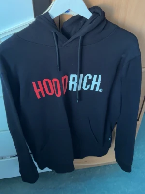 Svart HOODRICH hoodie med tryck - Snygg svart hoodie från HOODRICH med stor logga i rött och vitt på bröstet. Klassisk huva med dragsnören och en stor magficka framtill. Perfekt för en avslappnad streetwear-look. Tillverkad i mjukt material för extra komfort.