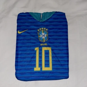 Blå Brasilien fotbollströja Nike - Snygg blå fotbollströja från Nike med Brasilien-emblem och nummer 10 i gult på bröstet. Tröjan har korta ärmar, gul Nike-logga och vågigt mönster över hela tyget. Perfekt för dig som älskar fotboll och vill sticka ut på planen.
