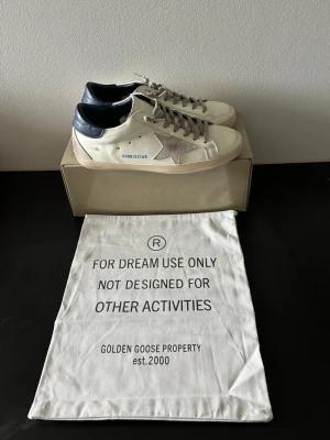 Golden Goose Superstar sneakers - Snygga Golden Goose Superstar sneakers i vitt läder med beige stjärndetalj på sidan och mörkblå häl. Klassisk låg modell med grå skosnören och slitna detaljer för en cool vintage-look. Tillverkade i Italien och levereras med dustbag.