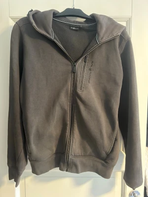 Mörkgrå hoodie från Sail Racing - Säljer en skön mörkgrå hoodie med dragkedja från Sail Racing i storlek medium. Den är i bra skick, det enda som är lite slitet är metall detaljerna på sidan av luvan. Kan gå ner i pris vid snabb affär.
