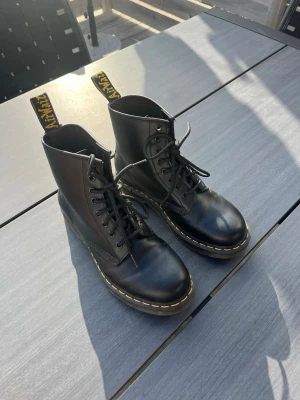 Svarta Dr. Martens skor - Klassiska svarta Dr. Martens skor i läder med gul söm runt sulan och högt skaft. Skorna har snörning framtill. Använda bara några få gånger så skicket är väldigt bra! Perfekta för dig som vill ha ett par nya lite finare skor. Nypris: 1750kr