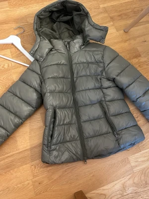 Grå pufferjacka med huva från 157 - Säljer en grå pufferjacka från 157 i storlek XS. Jackan har en avtagbar huva, två dragkedjeförsedda fickor och quiltad design. Perfekt för kalla dagar och riktigt skön att bära. Materialet är glansigt och vattenavvisande.