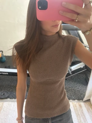 Ribbad brun ärmlös topp - Säljer en ribbad brun topp med hög hals och ärmlös design. Toppen har en tight passform och är gjord i ett mjukt stickat material som sitter snyggt på kroppen. Perfekt att styla med jeans eller kjol för en clean look.