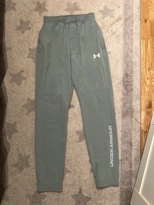 Gröna träningsbyxor Under Armour - Snygga gröna träningsbyxor från Under Armour med logga på låret och text längs benet. Byxorna har resår i midjan och vid bensluten samt praktiska sidofickor. Perfekta för gymmet eller löprundan.
