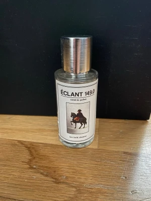 Éclant 149.0 extrait de parfum - Parfymflaska i glas med metallock och etikett med motiv av en ryttare. Flaskan har en stilren, cylindrisk form och etiketten är vit med svart text. Doften heter Éclant 149.0 och är märkt som extrait de parfum.