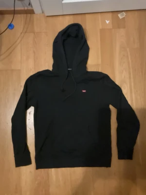 Svart Levi's Hoodie - Storlek M (Mycket bra skick) - Säljer en riktigt snygg och stilren svart hoodie från Levi's i storlek Medium (Standard fit). Tröjan är tillverkad i ett skönt och mjukt material med en klassisk passform som sitter riktigt bra. Den har den stilrena lilla röda Levi's-loggan placerad på bröstet, en rymlig känguruficka framtill samt huva med dragsko. Skicket är mycket bra då den bara är använd cirka 10 gånger. Den har inga hål, repor, trasiga sömmar eller andra skador. Perfekt basplagg till garderoben som passar till det mesta! 