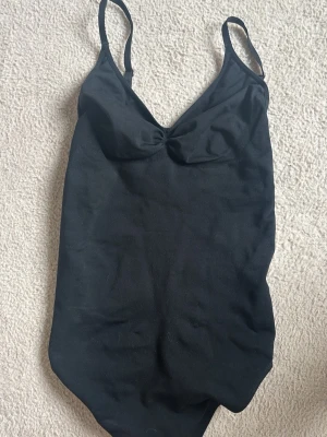 Svart enkel body med smala band - Snygg svart body med smala axelband och lätt rynkning vid bysten. Klassisk v-ringad modell som ger en stilren look. Perfekt för att få en fin aoutfit eller att ha under en annan tröja. Materialet känns stretchigt och bekvämt.Köpt för 200 kr