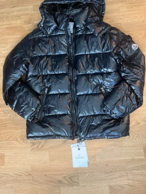 Svart dunjacka från Moncler - Snygg svart dunjacka från Moncler med glansig finish och klassisk puffer-design. Jackan har huva, två dragkedjeförsedda fickor och Moncler-logga på ärmen. Perfekt för kalla vinterdagar och riktigt trendig look.