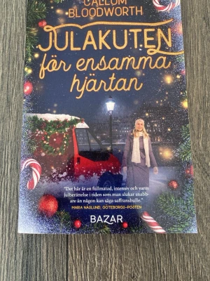 Julakuten för ensamma hjärtan - En mysig och varm julroman av Callum Bloodworth. Boken har ett färgstarkt omslag med juldekorationer, snö och en röd bil. Perfekt för dig som gillar feelgood, vänskap och romantik i vintermiljö. Utgiven av Bazar förlag.