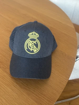 Mörkgrå Real Madrid keps - Snygg mörkgrå keps med Real Madrid-logga i gult framtill. Kepsen har böjd skärm och ventilerande mesh på sidorna för extra komfort. Perfekt för dig som vill visa ditt stöd för laget på ett stilrent sätt.