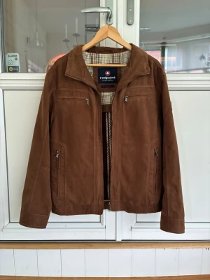 Brun skinnjacka från Redpoint - Snygg brun skinnjacka från Redpoint med klassisk krage och dragkedja framtill. Jackan har fyra fickor med dragkedja och rutigt innerfoder. Perfekt för dig som vill ha en stilren och tidlös look.
