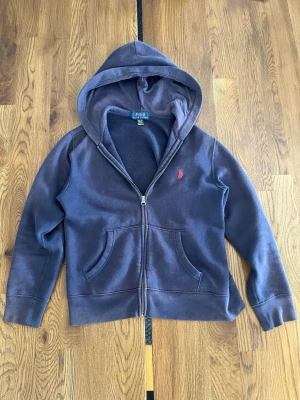 Polo Ralph Lauren - Mörkblå hoodie med dragkedja från Polo Ralph Lauren. Klassisk design med röd broderad logga på bröstet, två fickor fram och mjukt fleecefoder på insidan. Perfekt för en avslappnad och stilren look.