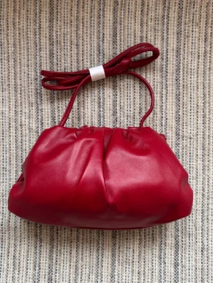 Röd clutchväska - Trendig röd handväska i mjukt material med puffig form och veckad design. Väskan har ett smalt axelband som kan bäras crossbody eller på axeln. Perfekt accessoar för att lyfta din outfit med en färgklick.
