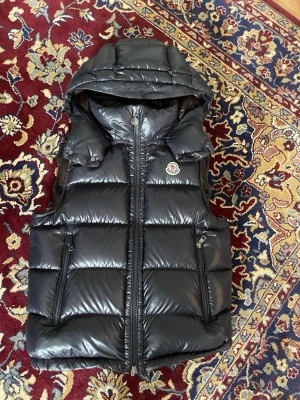 Moncler bormes väst  - KVITTO OCH TAGS FRÅN NK TILLKOMMER. Snygg svart dunväst från Moncler med hög krage och stor huva. Västen har blanka paneler, dragkedja framtill och Moncler-logga på bröstet. Perfekt för lager-på-lager och ger en clean streetwear-look. STORLEK 2