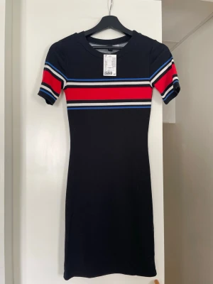 Ny med prislappen kvar svart klänning med röda, blå och vita detaljer - Helt ny med prislappen kvar, svart tight klänning från H&M i storlek 34. Jag är 173 och den går till mitten av låren på mig. Nypris 149kr.