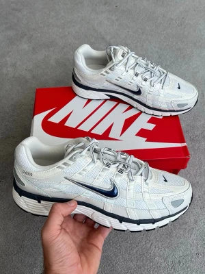 Nike P-6000 vita sneakers - Nike P-6000 sneakers i vitt med silver- och marinblå detaljer. Skorna har en sportig design med mesh och syntetmaterial, samt en tjock sula för extra komfort. Swoosh-logga på sidan och klassisk snörning framtill.