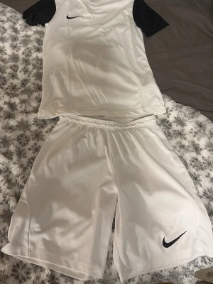 Vitt Nike träningsset med svarta detaljer - Säljer ett vitt träningsset från Nike med svart logga. Setet består av en t-shirt med svarta ärmar och ett par matchande vita shorts med resår i midjan. Perfekt för fotboll eller annan träning. Materialet är lätt och andas bra.