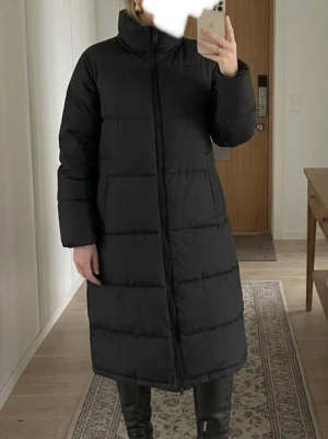 Svart lång dunjacka med dragkedja - Säljer en svart lång dunjacka med rak passform och diskreta horisontella sömmar. Jackan har hög krage, dragkedja framtill och två sidofickor. Perfekt för kalla vinterdagar och ger en stilren look.