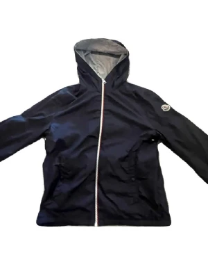 Svart vindjacka med huva från Moncler - Snygg svart vindjacka från Moncler med huva och vit dragkedja framtill. Jackan har två fickor och en Moncler-logga på ärmen. Tillverkad i lätt och tåligt material som skyddar mot vind. Perfekt för dig som vill ha en stilren och sportig look. STORLEK 14 år Men passar typ 13 år