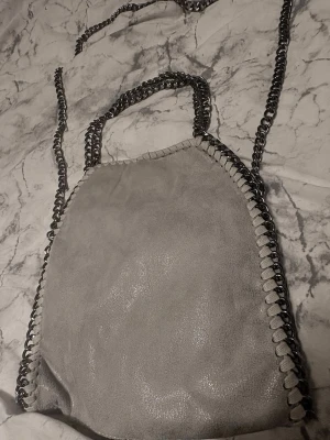 Grå axelväska med kedjedetaljer - Snygg grå axelväska i mjukt material med silverfärgade kedjor som axelrem och runt kanterna. Väskan har magnetknäppning och ett coolt innerfoder med leopardmönster. Perfekt för dig som gillar en edgy och trendig stil.