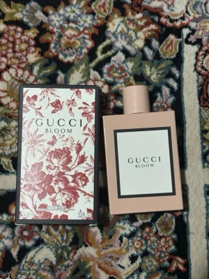 Gucci Bloom Eau de Parfum 100ml - Gucci Bloom Eau de Parfum i en stilren, ljusrosa rektangulär flaska med vit etikett och svart ram. Förpackningen har ett elegant blommönster i rött och vitt. Doften är känd för sina blommiga och fräscha noter.