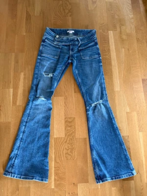 Blå bootcut jeans med slitningar - Säljer ett par blå bootcut jeans från Gina tricot i storlek XS. Jeansen har slitningar vid knäna och låret, samt stora fickor fram. Klassisk blå tvätt och utsvängda ben ger en retro vibe. Perfekta för dig som gillar en avslappnad men trendig stil. Knappt använd 