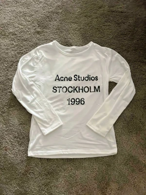 Vit långärmad topp Acne Studios - Vit långärmad topp från Acne Studios med tryck i svart text på bröstet där det står 'Acne Studios STOCKHOLM 1996'. Klassisk rund halsringning och mjukt material som känns skönt mot huden.