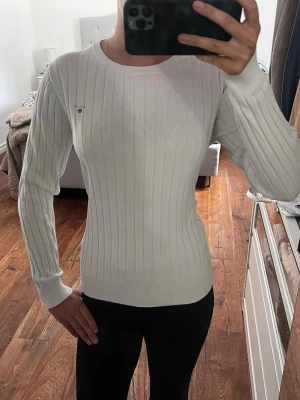 Vit ribbstickad tröja från Gant - Säljer en vit ribbstickad tröja från Gant med rund halsringning och diskret logga broderad på bröstet. Tröjan har långa ärmar och är tillverkad i mjukt material som sitter snyggt på kroppen. Perfekt för en clean och klassisk look.