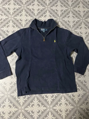 Mörkblå halvzip från Polo Ralph Lauren - Klassisk mörkblå halvzip-tröja från Polo Ralph Lauren med gul broderad logga på bröstet. Tröjan har hög krage, dragkedja med läderdetalj och långa ärmar. Perfekt för en avslappnad men ändå stilren look.