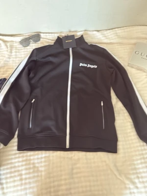 Svart track jacket  - Säljer en svart track jacket med vita detaljer längs dragkedjan och ärmarna. Jackan har hög krage, två fickor med dragkedja. Perfekt för en sportig och trendig look.