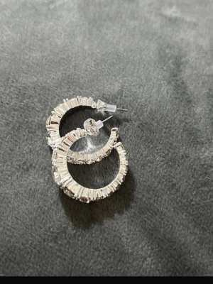 Silvriga creolörhängen med stenar - Säljer ett par silvriga creolörhängen med gnistrande, rektangulära stenar runt hela ringen. Örhängena har en halvcirkelformad design och är tillverkade i metall med infattade klara stenar som ger en lyxig känsla.