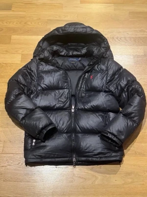 Ralph Lauren jacka  - Säljer en svart pufferjacka med huva och dragkedja framtill. Jackan har en klassisk quiltad design, två sidofickor med dragkedja och en liten röd logga på bröstet. Har lagats hos veriferad skräddare hålet på jackan. 