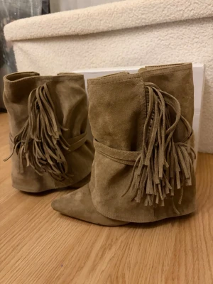 Beige boots med fransar i mocka - Säljer ett par beige boots i mjuk mocka med dekorativa fransar på sidan. Bootsens design är slouchy med rundad tå och låg klack, vilket ger en avslappnad men trendig look. Perfekta för dig som gillar boho-stil och vill sticka ut.