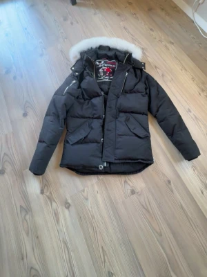 Moose knuckles jacka - Snygg svart dunjacka från moose knuckles med avtagbar huva och vit pälskant. Jackan har flera fickor, dragkedja och tryckknappar framtill samt detaljerad patch på insidan. Perfekt för kalla vinterdagar och riktigt varm. Varan är äkta! Och tillkommer med kvitto och väskan man får med när man köper den. Den är i storlek s men skulle säga att den passar m lika bra! Skriv för mer bilder jackan är i väldigt bra skick!