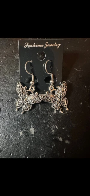 Silvriga fjärilsörhängen - Unika örhängen i silverfärgad metall med detaljerade fjärilar. Hängande modell med krok, perfekt för dig som gillar statement-smycken och vill addera en cool touch till din stil. Fjärilarna har fina mönster och ger ett elegant intryck.