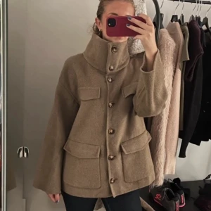 Beige ulljacka med stora fickor - Snygg beige jacka i ullmix med stora knappar och fyra rejäla fickor framtill. Jackan har hög krage som kan bäras uppvikt eller nedvikt och en rak, något boxig passform. Perfekt för lager på lager och ger en trendig look till hösten.