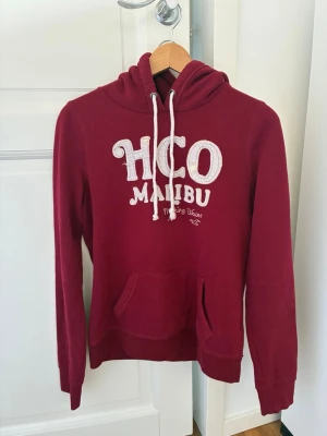 Vinröd HCO Malibu hoodie med huva - Snygg vinröd hoodie i märket Hollister, med stor vit HCO Malibu-text på bröstet och broderad detalj. Klassisk känguruficka framtill, långa ärmar och justerbar huva med vita snören. Perfekt för en avslappnad och trendig look. Storlek L, upplevs mer som S. 