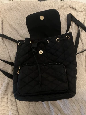Liten svart quiltad ryggsäck med snörning - Snygg svart quiltad ryggsäck med snörning och magnetlås. Väskan har justerbara axelband, en ficka framtill med dragkedja och guldfärgade detaljer. Perfekt för dig som vill ha en stilren och praktisk accessoar till skolan eller stan. Liten storlek. 