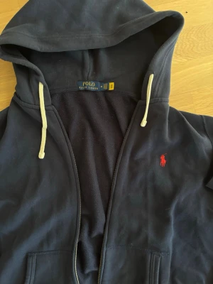 Ralph lauren zip hoodie - Snygg marinblå hoodie med dragkedja från Polo Ralph Lauren. Klassisk design med vit dragsko i huvan och röd broderad logga på bröstet. Mjuk bomullskänsla och två fickor framtill. Perfekt för en avslappnad och stilren look.