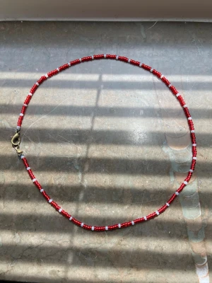 Rött och vitt pärlhalsband - Säljer ett handgjort halsband med små röda och vita glaspärlor. Halsbandet har ett enkelt och stilrent mönster med metallfärgat lås. Perfekt för dig som gillar färgglada accessoarer och vill addera något unikt till din stil.