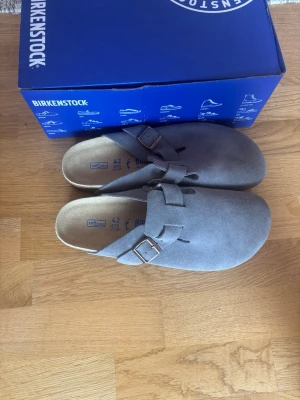 Grå Birkenstock Boston - Säljer ett par grå Birkenstock Boston tofflor i mocka med justerbart spänne i metall. Klassisk design med mjuk fotbädd och öppen häl. Perfekta för chill dagar hemma eller när du vill ha det bekvämt.