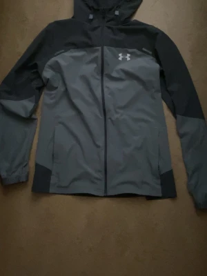 Grå och svart vindjacka Under Armour - Snygg vindjacka från Under Armour i grått och svart med huva och dragkedja framtill. Jackan har logga på bröstet och text på ryggen. Tillverkad i lätt och tåligt material, perfekt för blåsiga dagar. Två sidofickor och elastiska muddar vid ärmsluten.