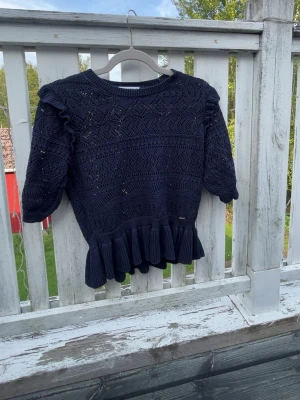 Mörkblå stickad topp med volang - Snygg mörkblå stickad topp med korta ärmar och volangkant nedtill. Tröjan har ett detaljerat hålmönster och ribbad halsringning. Perfekt för dig som gillar en feminin och trendig stil.💙