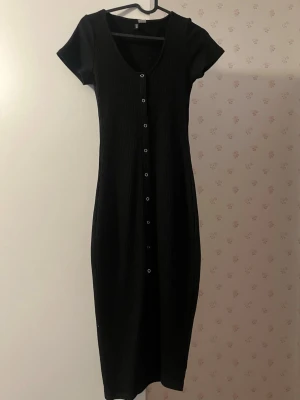 Svart ribbad klänning med knappar - Svart ribbad klänning med korta ärmar och v-ringning. Klänningen har knappar längs hela framsidan och är i en längre modell. Perfekt för dig som gillar enkel men stilren design. Oanvänd 🫶 