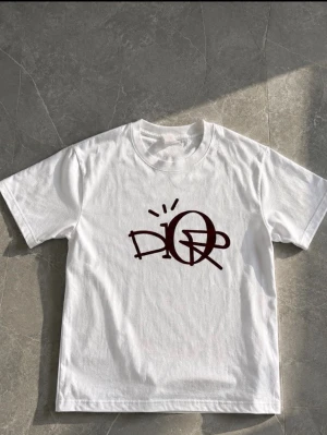 Vit t-shirt från dior i  vinrött  - Säljer en vit t-shirt med ett stort vinrött grafiskt tryck på bröstet. T-shirten har klassisk passform, rund halsringning och korta ärmar. Perfekt för dig som gillar streetwear och enkel stil.